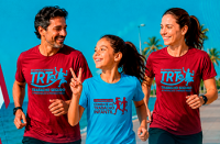 Há um homem, uma mulher e uma menina correndo na praia com camisetas do TRT 