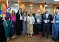 A imagem mostra um grupo de oito pessoas — seis mulheres e dois homens — alinhadas lado a lado em uma sala institucional decorada com painéis de madeira e um crucifixo ao fundo. Todos vestem trajes formais ou esporte fino, e quase todas as pessoas seguram um exemplar da mesma cartilha com o título "Violência Doméstica". As mulheres vestem roupas em tons variados de verde, rosa, preto, bege e cinza, enquanto os homens à direita usam terno e gravata. No lado esquerdo, é possível ver as bandeiras do Brasil.