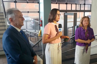 A imagem mostra três pessoas em um ambiente interno. Ao centro, uma mulher segura um microfone durante uma entrevista, enquanto um homem, à esquerda, e outra mulher, à direita, acompanham a gravação. Ao fundo, há grandes portas de vidro.