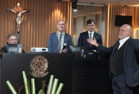 A imagem mostra o Plenário do TRT-19 em tom de celebração, com o Brasão da República em destaque na bancada e um crucifixo de madeira ao fundo. Um homem de terno escuro, colete e barba branca sorri de braços abertos em primeiro plano, demonstrando entusiasmo para a plateia. Atrás da bancada, três autoridades em trajes formais acompanham a cena sorridentes, uma delas usando um colar colorido e segurando um microfone.