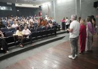 A imagem captura um evento em um auditório, onde um homem de camiseta branca fala ao microfone para uma plateia atenta, tendo ao seu lado três mulheres com vestes coloridas. O público está sentado em poltronas azuis dispostas em fileiras escalonadas, ocupando a maior parte do espaço e voltado para os palestrantes. O ambiente tem paredes de concreto exposto e teto com iluminação embutida e saídas de ar condicionado. No palco, destaca-se o piso de madeira rústica, onde o grupo de oradores está posicionado. 