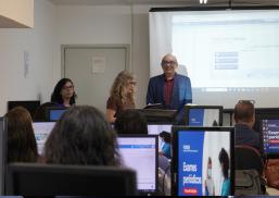 HÁ DUAS MULHERES E UM HOMEM DE PÉ FALANDO PARA VÁRIAS PESSOAS SENTADAS EM UMA SALA DE AULA, COM COMPUTADORES À SUA FRENTE.