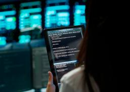 Uma pessoa é vista de costas segurando um tablet que exibe uma tela com código de programação. Ao fundo, há monitores desfocados com interfaces digitais em tons de azul e verde, compondo um ambiente tecnológico que remete a um centro de processamento de dados ou sala de monitoramento de sistemas.