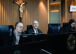 Uma sessão de julgamento no plenário do TRT-AL, com três magistrados sentados à bancada. Ao centro, o desembargador Antônio Catão sorri para a câmera, enquanto os demais participantes acompanham os trabalhos. Ao fundo, destaca-se um crucifixo fixado na parede revestida em madeira e um monitor indicando o horário da sessão.