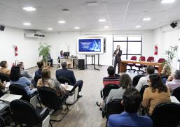 Auditório com várias pessoas sentadas. Elas assitem uma pessoa falar. Há um telão atrás da mulher que fala.