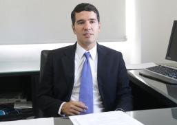 Há um homem de terno preto e gravata lilás, sentado e trabalhando em uma mesa com computador