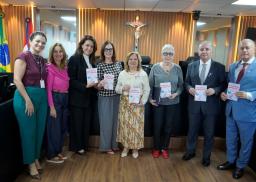 A imagem mostra um grupo de oito pessoas — seis mulheres e dois homens — alinhadas lado a lado em uma sala institucional decorada com painéis de madeira e um crucifixo ao fundo. Todos vestem trajes formais ou esporte fino, e quase todas as pessoas seguram um exemplar da mesma cartilha com o título "Violência Doméstica". As mulheres vestem roupas em tons variados de verde, rosa, preto, bege e cinza, enquanto os homens à direita usam terno e gravata. No lado esquerdo, é possível ver as bandeiras do Brasil.