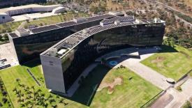 Esta fotografia aérea exibe o complexo arquitetônico do Tribunal Superior do Trabalho (TST) e do Conselho Superior da Justiça do Trabalho (CSJT), em Brasília.  Principais elementos da imagem:  Arquitetura: Dois edifícios modernos e curvos com fachadas em vidro escuro espelhado. Na extremidade de um dos blocos, destacam-se as siglas "TST" e "CSJT" gravadas em relevo na parede cinza.  Entorno e Paisagismo: Amplas áreas gramadas, palmeiras e vias de acesso cercam a estrutura.  