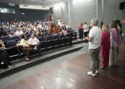 A imagem captura um evento em um auditório, onde um homem de camiseta branca fala ao microfone para uma plateia atenta, tendo ao seu lado três mulheres com vestes coloridas. O público está sentado em poltronas azuis dispostas em fileiras escalonadas, ocupando a maior parte do espaço e voltado para os palestrantes. O ambiente tem paredes de concreto exposto e teto com iluminação embutida e saídas de ar condicionado. No palco, destaca-se o piso de madeira rústica, onde o grupo de oradores está posicionado. 