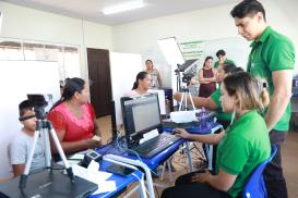 A imagem mostra uma ação de atendimento comunitário em uma sala bem iluminada, onde atendentes de camisa verde operam computadores para cadastrar cidadãos. Em primeiro plano, uma mulher de blusa vermelha e um garoto aguardam enquanto passam por um procedimento com câmera e leitor biométrico. Ao fundo, outra mulher está sentada diante de uma luz de iluminação (softbox) sendo fotografada ou atendida por outra funcionária. Diversas pessoas aguardam de pé e observam o processo, que conta com equipamentos de TI 