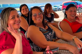 A imagem mostra um grupo de cinco mulheres sorridentes reunidas sob a sombra de uma estrutura aberta ou tenda. Em primeiro plano, uma mulher à esquerda, vestindo uma blusa vermelha, e outra à direita acenam amigavelmente para a câmera. Ao centro, outra mulher de óculos e blusa estampada sorri abertamente enquanto segura um celular. Ao fundo, mais duas mulheres completam a cena, também bastante sorridentes e descontraídas. A iluminação é natural e luminosa, revelando um estacionamento com diversos carros