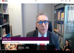 A imagem mostra o presidente do TRT da 19ª Região, desembargador Jasiel Ivo, em uma entrevista por videoconferência. Ele aparece de terno e gravata, diante de uma estante de livros, enquanto uma intérprete de Libras realiza a tradução no canto inferior direito da tela.