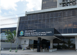 A foto exibe a fachada do prédio do Fórum Quintella Cavalcanti, onde funcionam as Varas do Trabalho de Maceió. O edifício possui estrutura moderna com amplas janelas espelhadas de tom escuro e a identificação oficial na entrada. Na parte inferior, vê-se a recepção coberta, bancos de espera na calçada e uma área demarcada por fitas zebradas.